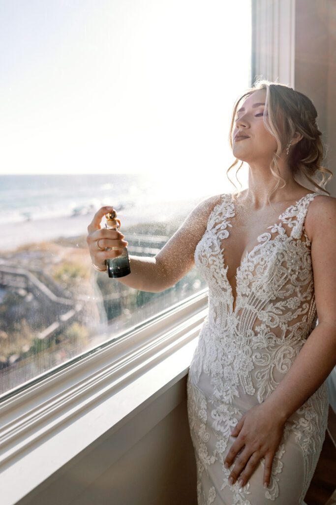 Bride spraying perfume on herself in front of large window overlooking the beach in Destin, FL