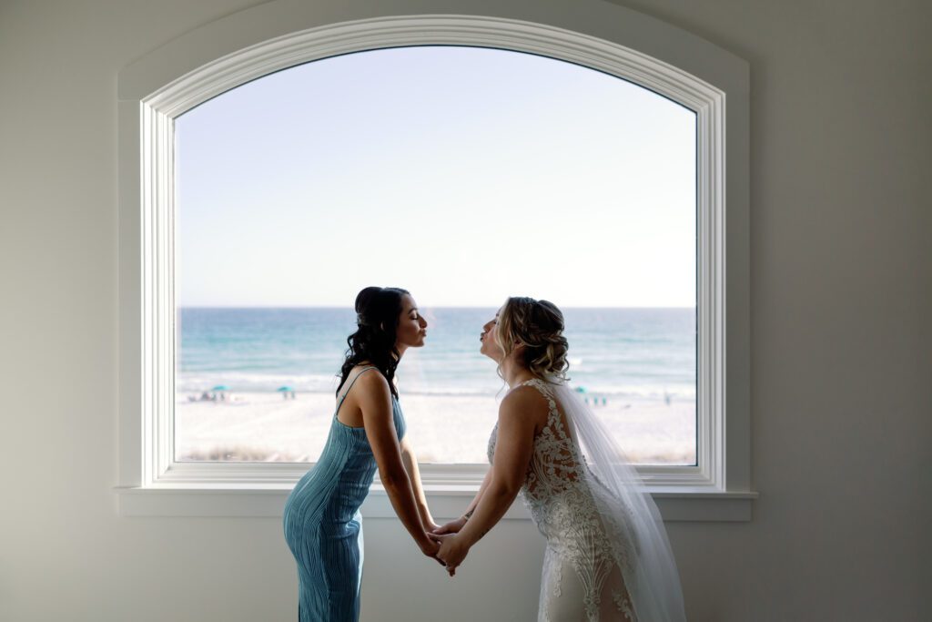 Maid of Honor and Bride blowing kisses at each other in front of a large window overlooking the beach in Destin, FL